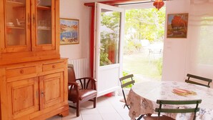 A dining area with a wooden cabinet, chairs, a table with a tablecloth, and a view of the garden through a glass door. - ARTIST'S HOME, 5 MINUTES FROM THE SEA, VERY QUIET, TUCKED AWAY IN A LUSH GREEN SETTING. (Trégastel)