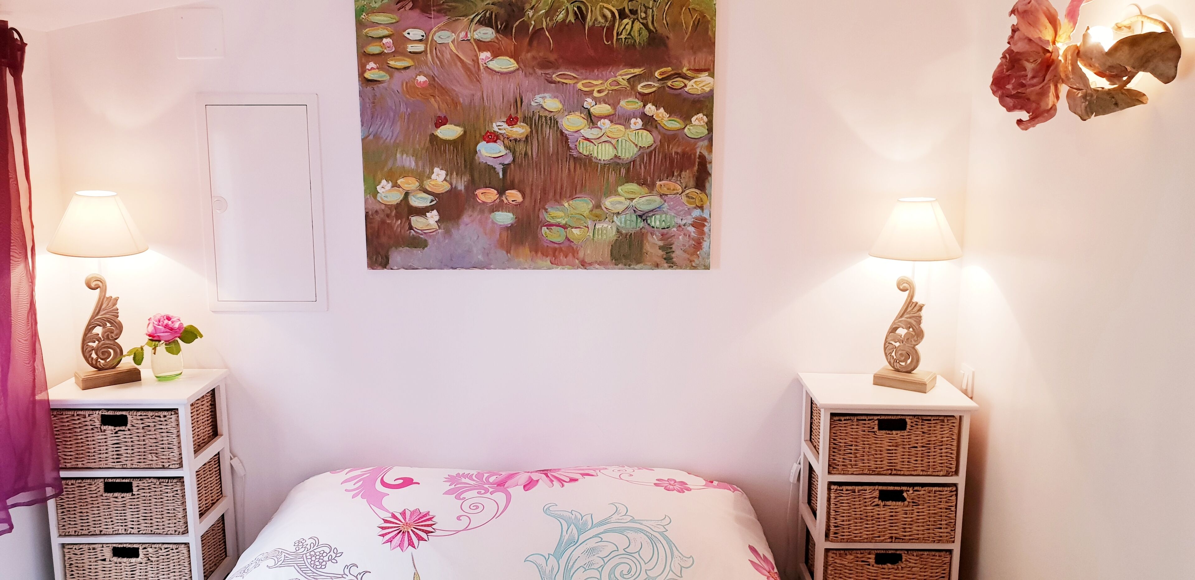 Un dormitorio con una cama, dos mesitas de noche con lámparas, cestas de mimbre, un cuadro y una puerta.