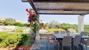 An outdoor patio with a table and chairs, surrounded by greenery and a pergola with red flowers.