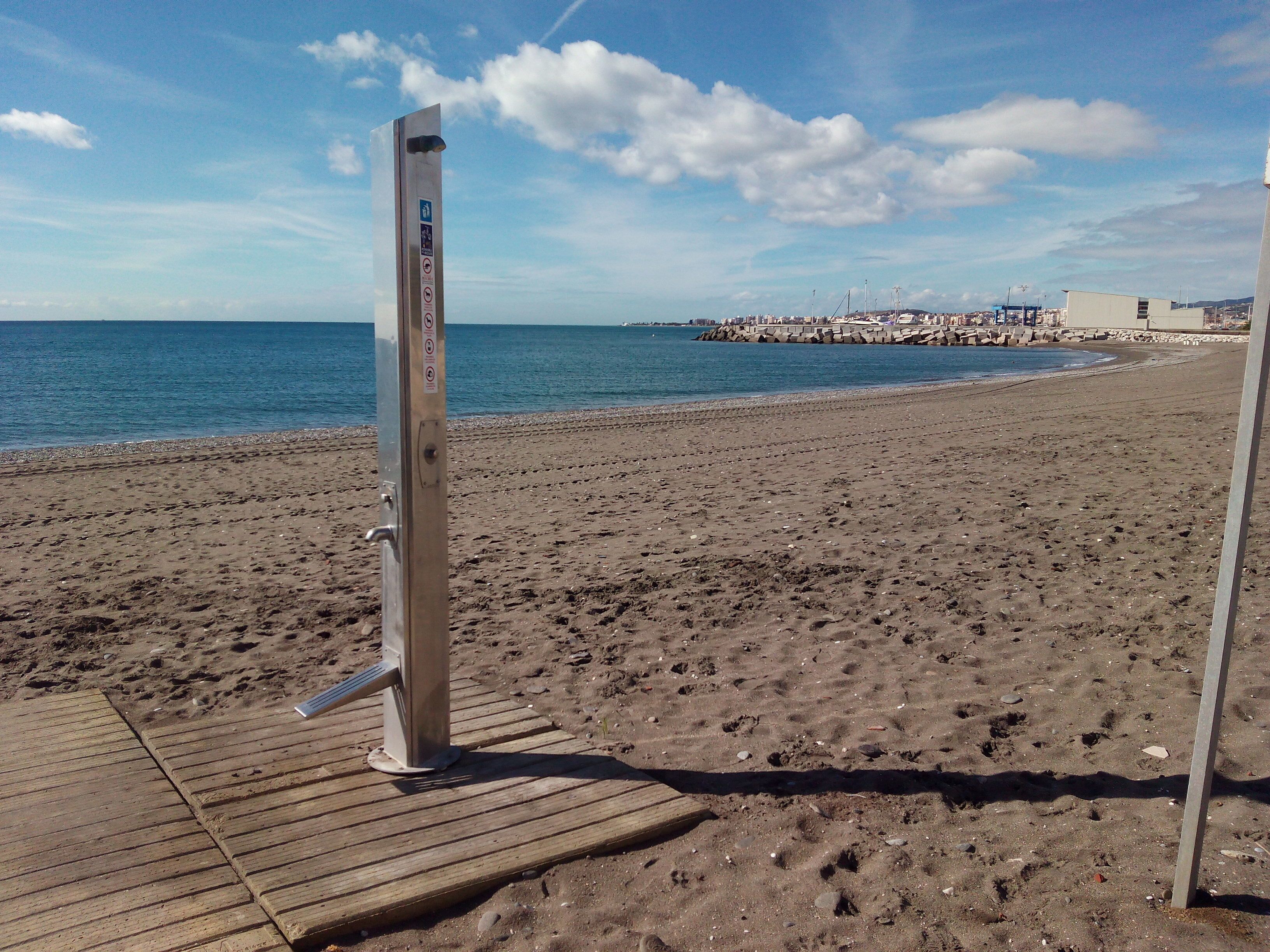 A beach with a wooden boardwalk, a metal structure with a sign, and a distant view of a pier with buildings.