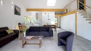 Un salón amplio con un sofá oscuro, una mesa de centro de madera y una silla moderna. Una escalera conduce a un nivel superior con barandilla de madera.