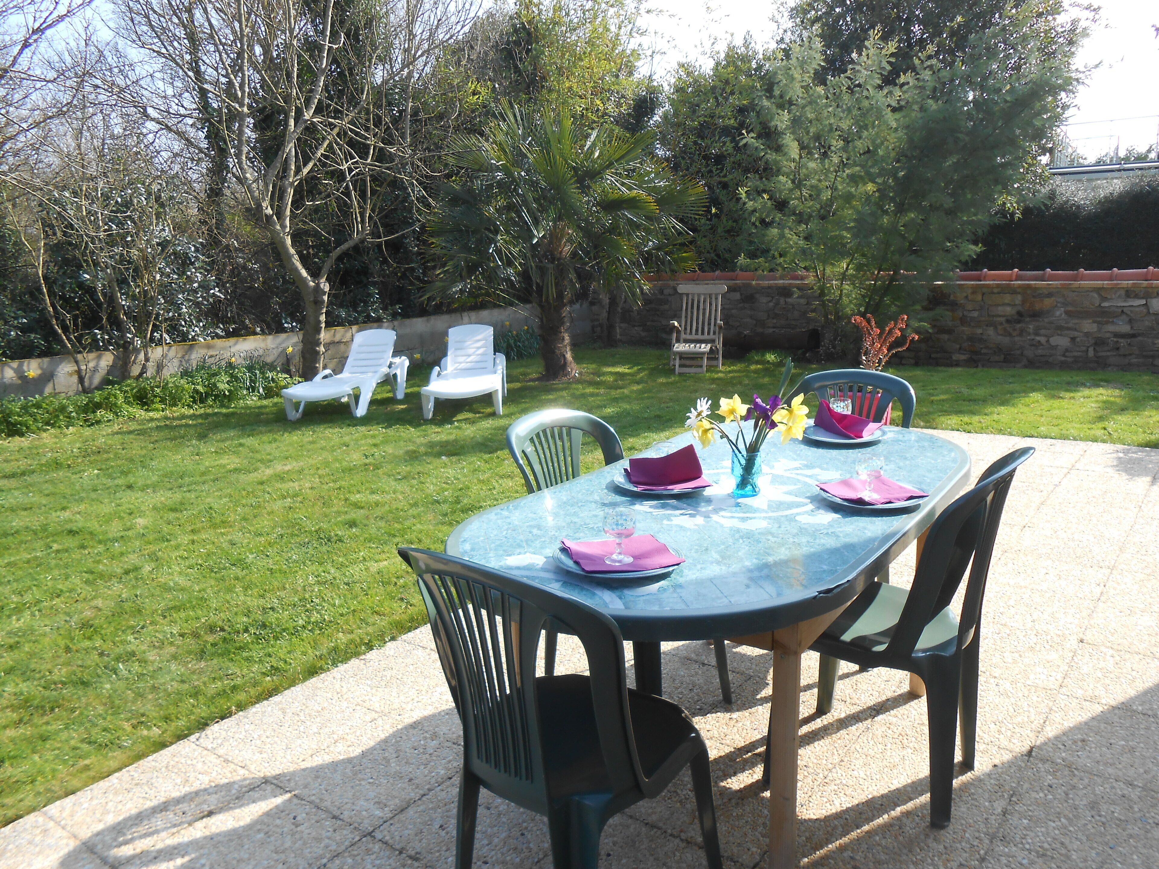 A patio with a table set for two, surrounded by chairs, a stone wall, and a grassy area with lounge chairs.