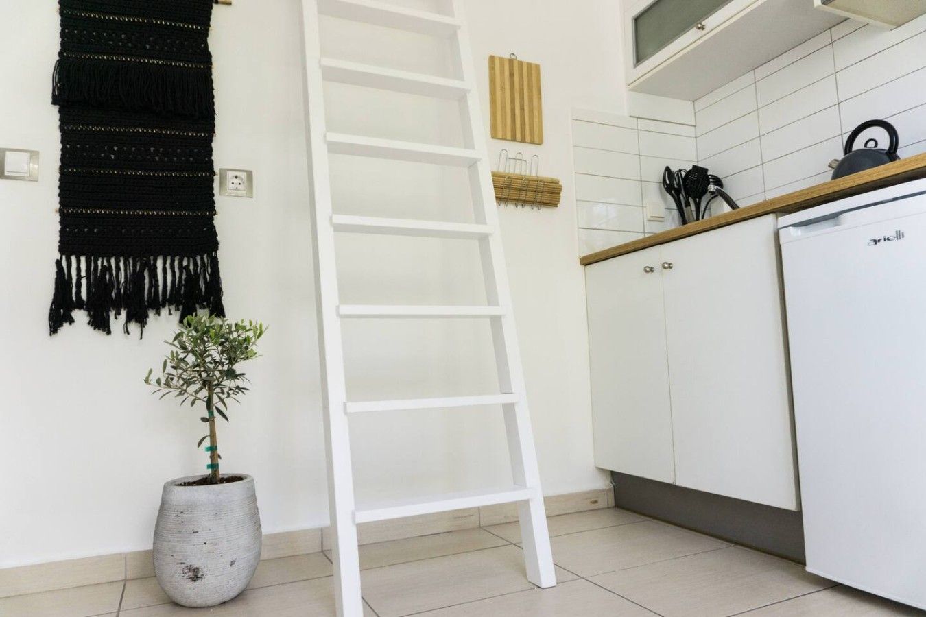 Une cuisine avec des meubles blancs, un réfrigérateur blanc, une décoration murale noire, une échelle blanche et une plante en pot.