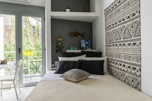A bedroom with a bed, pillows, a wall with a geometric pattern, a shelf with decorative items, and a view of greenery through a sliding door.