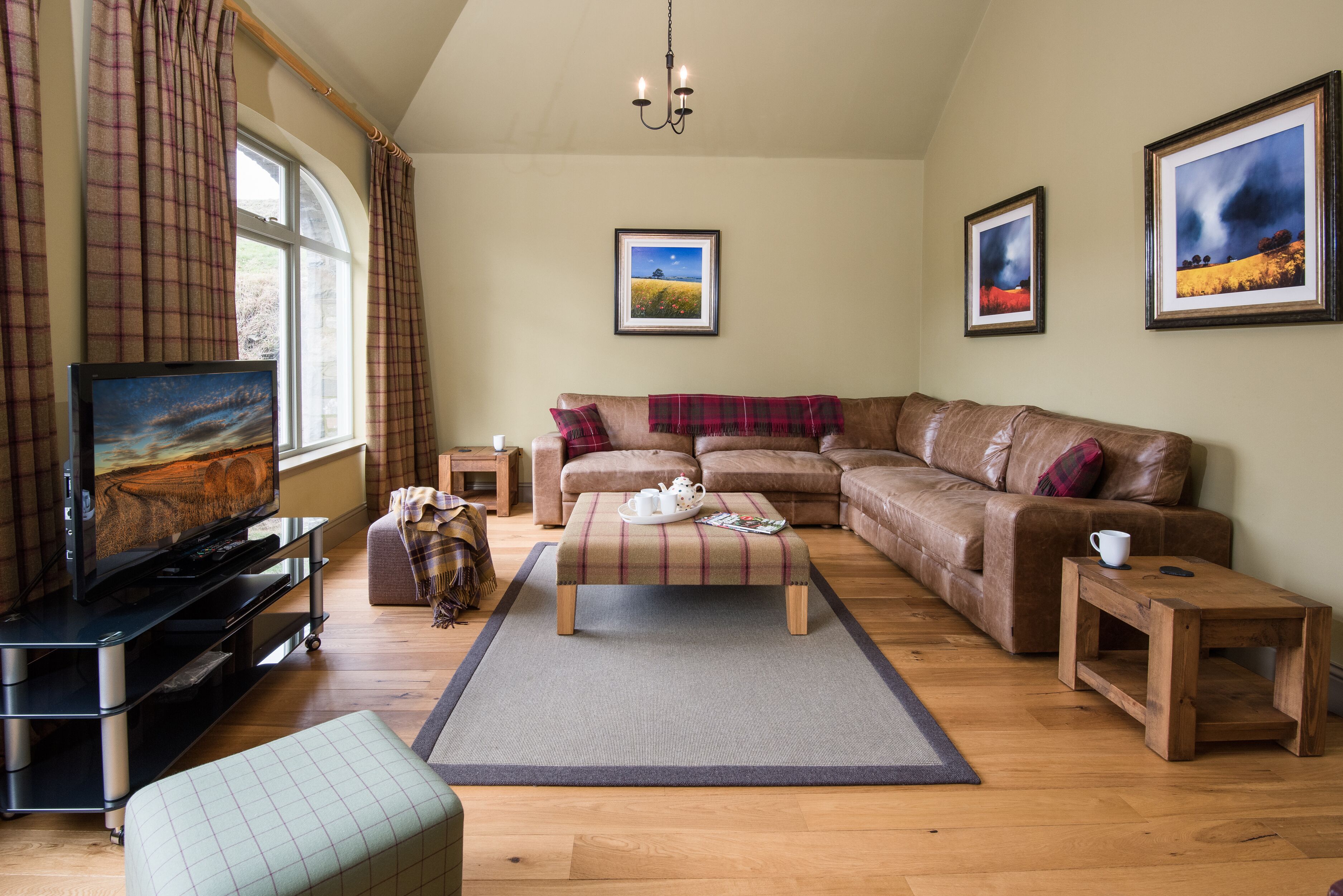 A cozy living room with a large window, a flat-screen TV, a brown leather sofa, a plaid ottoman, and a wooden coffee table.