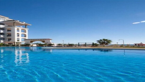 A swimming pool with a clear blue water surface, surrounded by a white building with multiple balconies and an archway entrance. - Residencial Marina Duquesa 642 (Manilva)