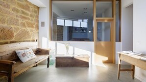 A room with a stone wall, a wooden bench with a cushion, a small wooden table, and a large window with a view of a kitchen.