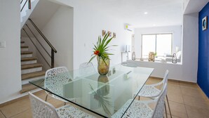A modern dining area with a glass table, white chairs, a vase with a plant, a staircase, and a view of a balcony with outdoor furniture. - Central Amazing PH with Private Rooftop and Jacuzzi (Playa del Carmen)