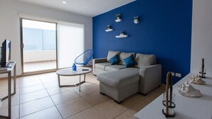 A modern living room with a grey sofa, a blue chair, and a coffee table. There are decorative plants on shelves and a large window with a view. - Central Amazing PH with Private Rooftop and Jacuzzi (Playa del Carmen)