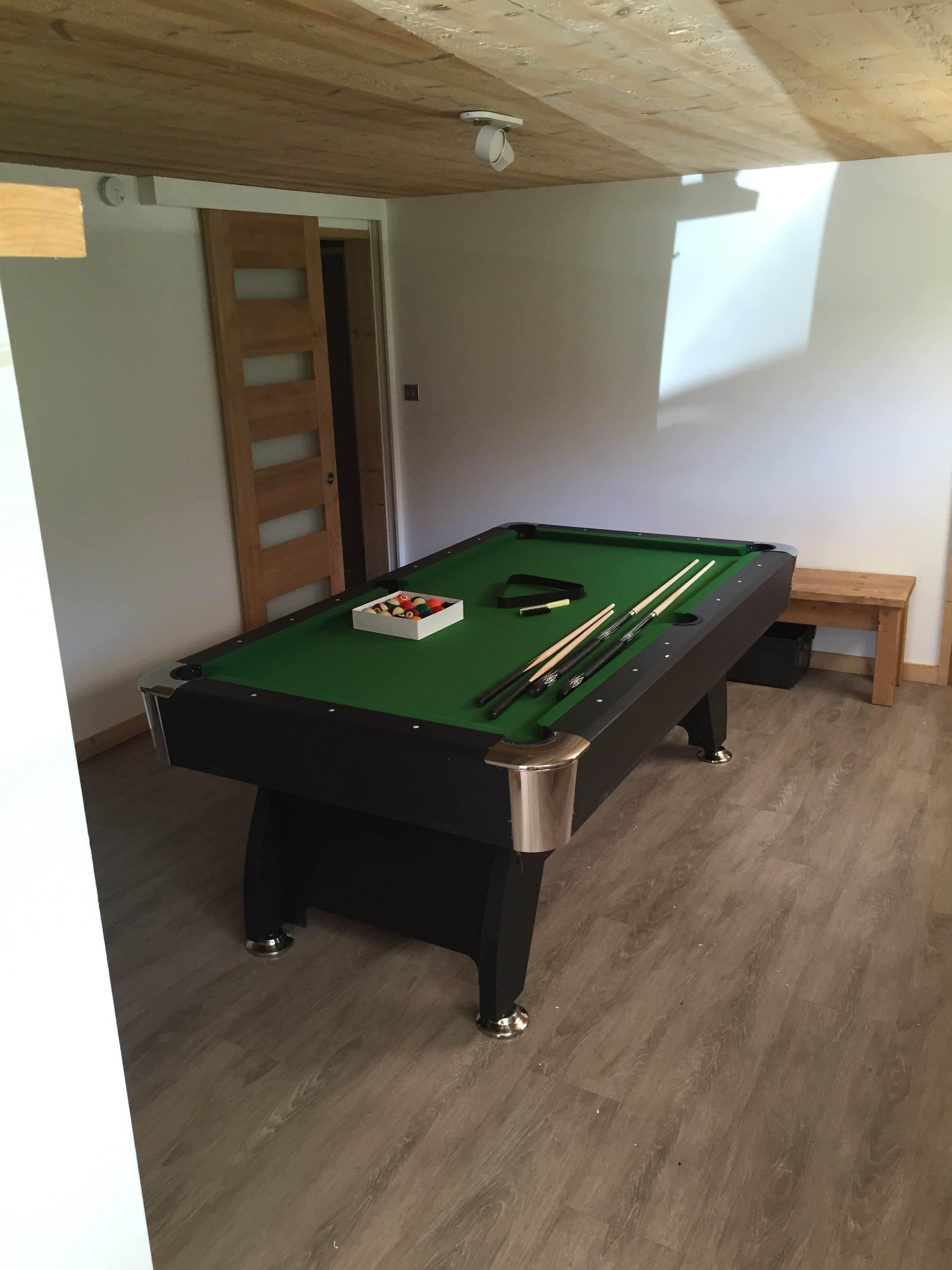 Una mesa de billar con bolas y tacos, un banco de madera y una puerta en una habitación con suelo de madera y techo inclinado.
