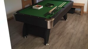 Une table de billard avec des billes et des queues, un banc en bois, et une porte dans une pièce dont le sol est en bois et dont le plafond est incliné.