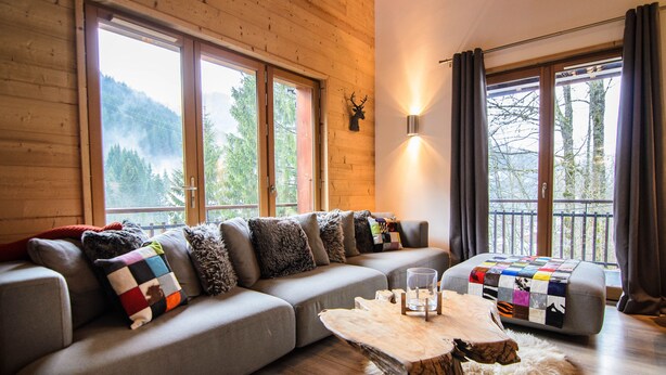 Un salon moderne avec un canapé gris, une table basse en bois et une vue sur l’extérieur grâce à de grandes fenêtres.