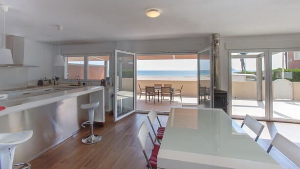 Een moderne keuken met een groot eiland, een eetgedeelte met een glazen tafel en uitzicht op het strand door grote schuifdeuren.
