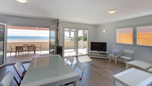 Een moderne woonkamer met een glazen deur die uitkomt op een balkon met uitzicht op een strand, een eethoek met stoelen, een televisie en een klein tafeltje.