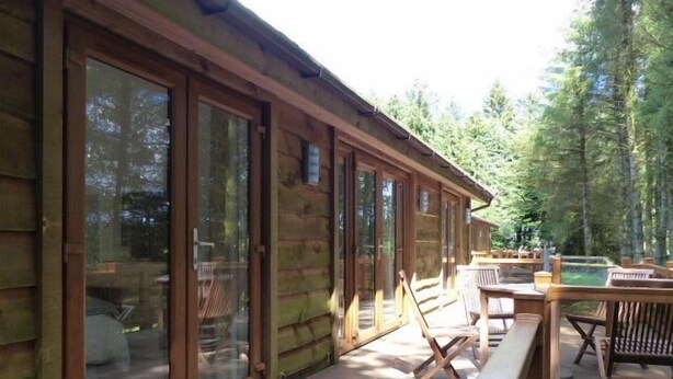 Eine Holzhütte mit großen Schiebeglastüren, einer Terrasse mit Holzmöbeln und einem bewaldeten Hintergrund.