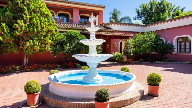 Une fontaine à plusieurs niveaux, entourée de plantes en pot, se dressant devant un bâtiment doté d’un toit en tuiles et de murs rouges.