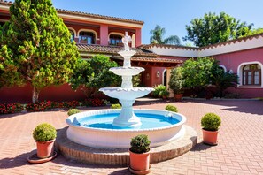 A fountain with a tiered design surrounded by potted plants, set against a building with a tiled roof and red walls. - Luxury villa in Marbella (Guadalmina Baja)  (Marbella)