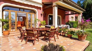 Une terrasse avec une table ronde et des chaises, entourée de plantes en pot et d’une pelouse.