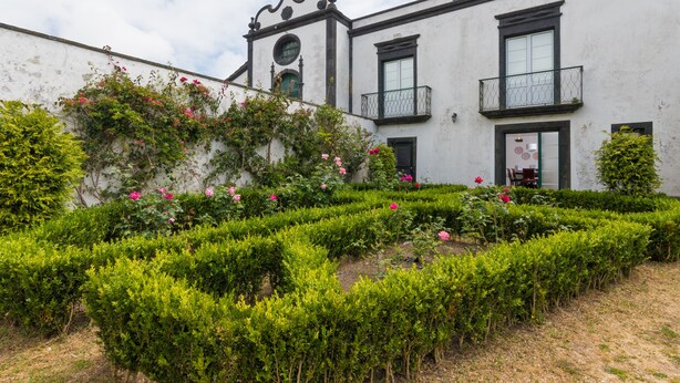 A building with a white facade, a bell tower, and a garden with hedges and flowers. - Casa do Loreto - Ponta Delgada (Fajã de Baixo)