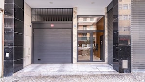A modern building entrance with a closed grey roller door, a visible fire alarm, and a glass door leading to a parking area.