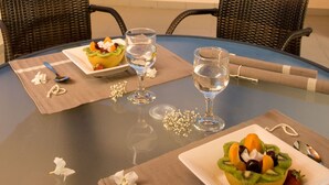 Et bord dækket med en skål frugt, to vinglas og en tallerken mad på en balkon med udsigt over havet.