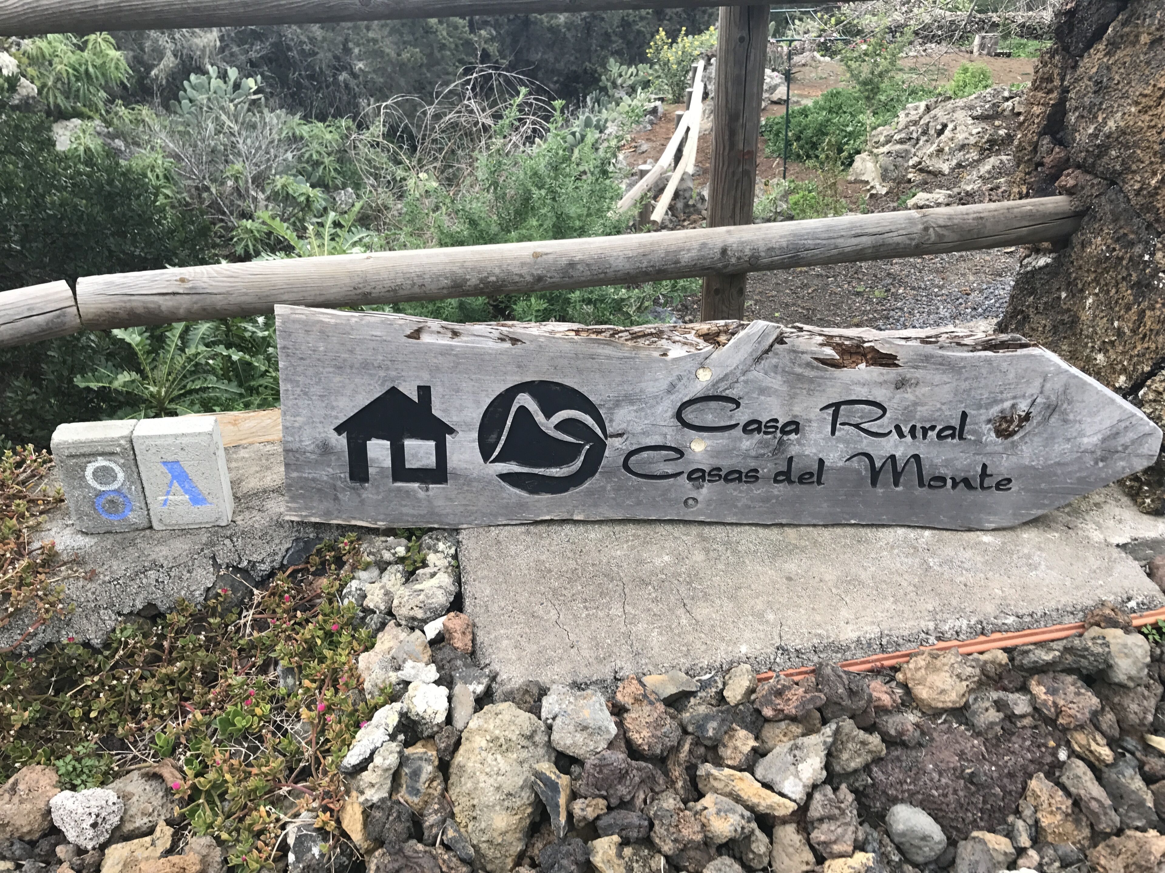 A rustic wooden sign with a house and mountain icon, indicating a rural lodging area named 'Casa Rural Casas del Monte'.