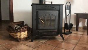 A black wood-burning stove with a glass door and a metal handle, placed on a tiled floor next to a wicker basket filled with pinecones. - Gîte CASAS DEL MONTE, your refuge in the middle of nature. (Valverde)