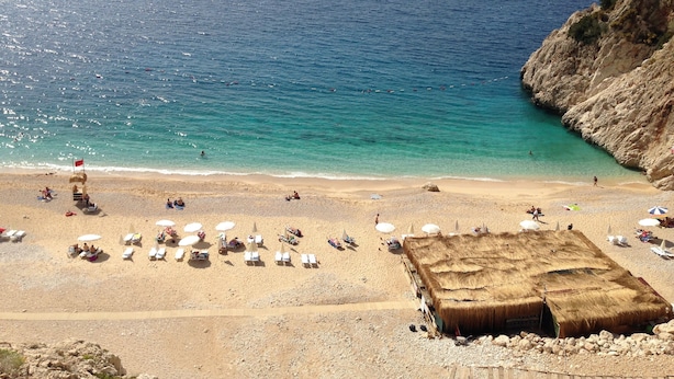 A beach with sun loungers and umbrellas, a thatched hut, and rocky cliffs. - Apartment With Shared Pool And Sea Views (Kalkan)
