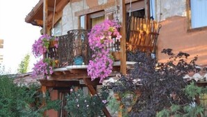 A rustic wooden house with a balcony adorned with pink flowers, surrounded by lush greenery.