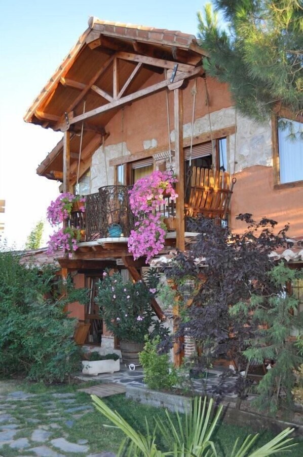 Une maison en bois à l’allure rustique, dotée d’un balcon orné de fleurs roses et entourée d’une végétation luxuriante.