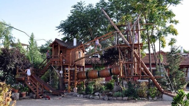 A wooden play structure with slides and climbing elements, surrounded by trees and potted plants.