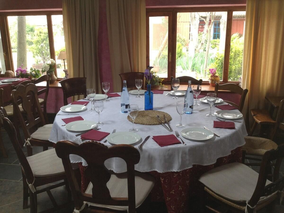 Une table de salle à manger ronde, mise pour un repas, entourée de chaises, avec un vase de fleurs et une vue sur la verdure à l’extérieur.
