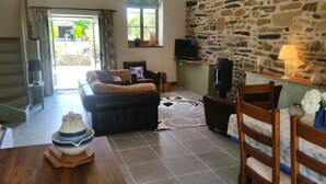 A living room with a stone wall, a television, a sofa, and a dining area with a table and chairs. - Beautiful, spacious, 2 bed renovated breton cider press, 10km from the coast! (Cast)