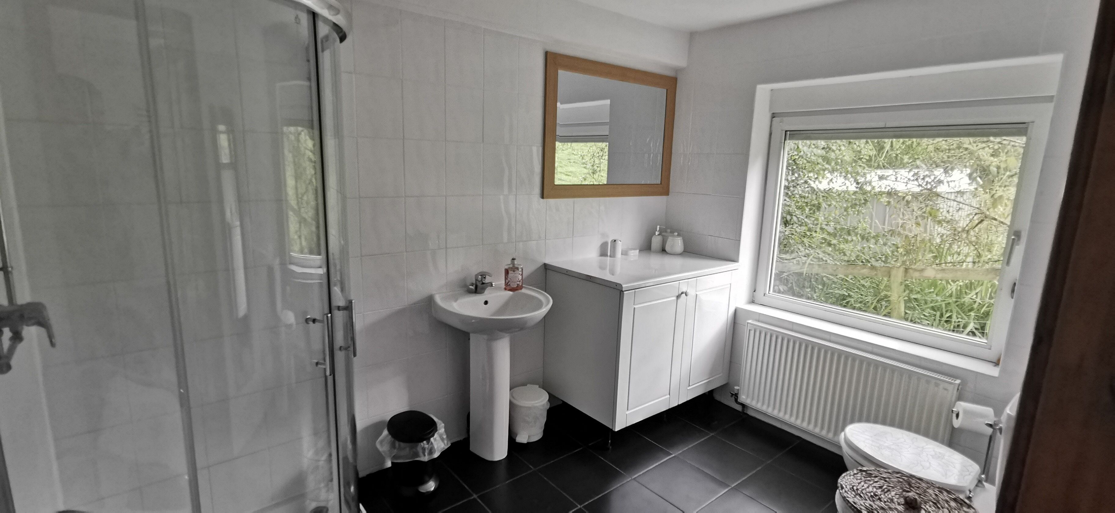 A modern bathroom with a glass shower, white tiles, a sink, a window, and a toilet.