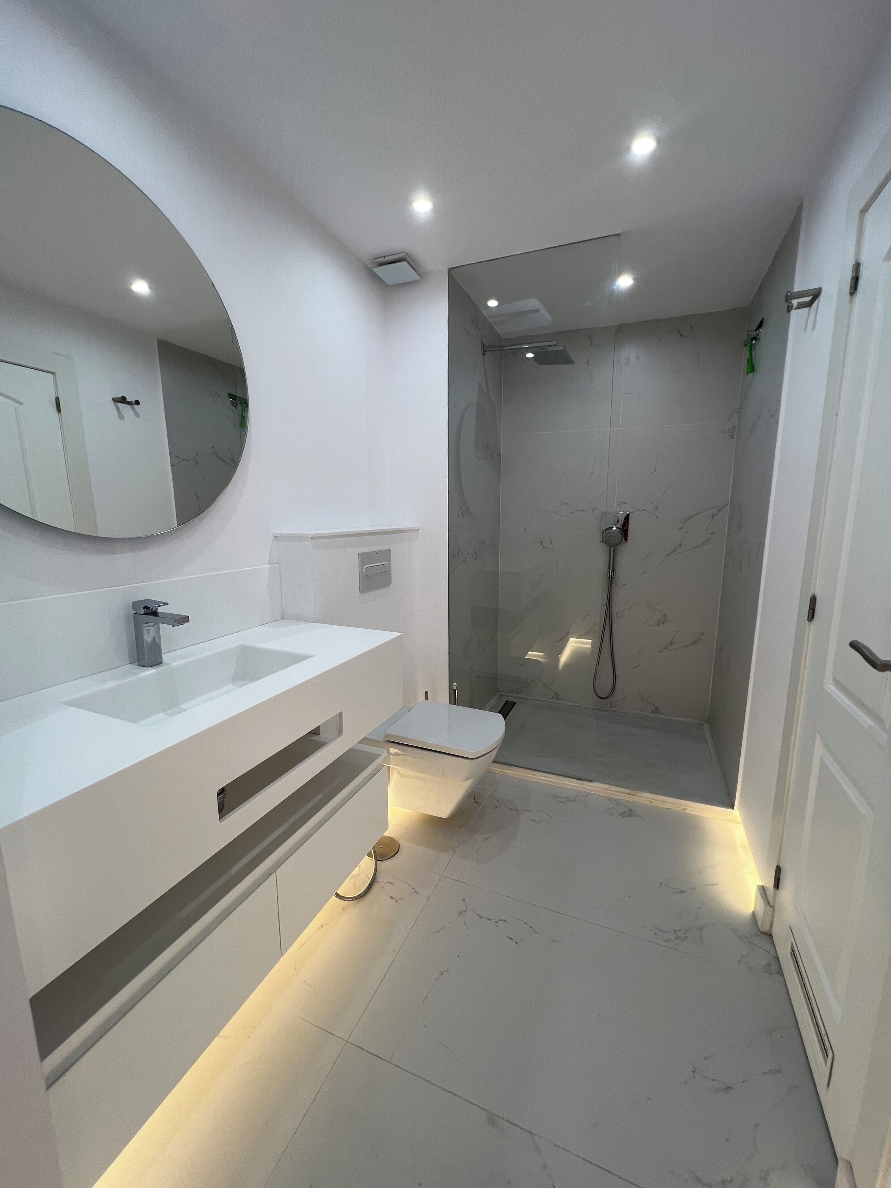 A modern bathroom with a white vanity, a round mirror, a shower with a glass door, and recessed ceiling lights.