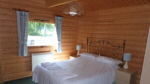 A wooden cabin bedroom with a bed, two bedside tables with lamps, a window with curtains, and a ceiling fan.