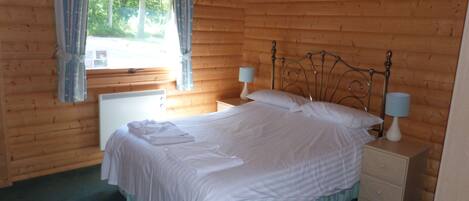 A wooden cabin bedroom with a bed, two bedside tables with lamps, a window with curtains, and a ceiling fan.