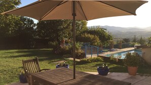 Eine Terrasse im Freien mit Holztisch und -stühlen, einem großen Sonnenschirm und Blick auf ein Schwimmbad und Berge im Hintergrund.
