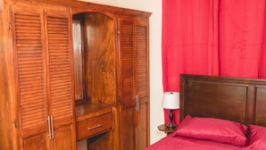 A bedroom with a wooden wardrobe, a bed with red bedding, and a bedside table with a lamp. - Frontline apartment No. 2 (Oistins)