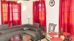 A living room with a grey sofa, a ceiling fan, red curtains, a dining table with chairs, and a clock on the wall. - Frontline apartment No. 2 (Oistins)