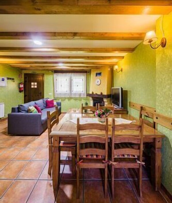 Un comedor con una mesa y sillas de madera, un sofá y un televisor en una habitación con paredes verdes y vigas de madera en el techo.