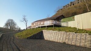 A building with a sloped roof and a balcony, situated on a hillside with a gravel driveway leading to it.