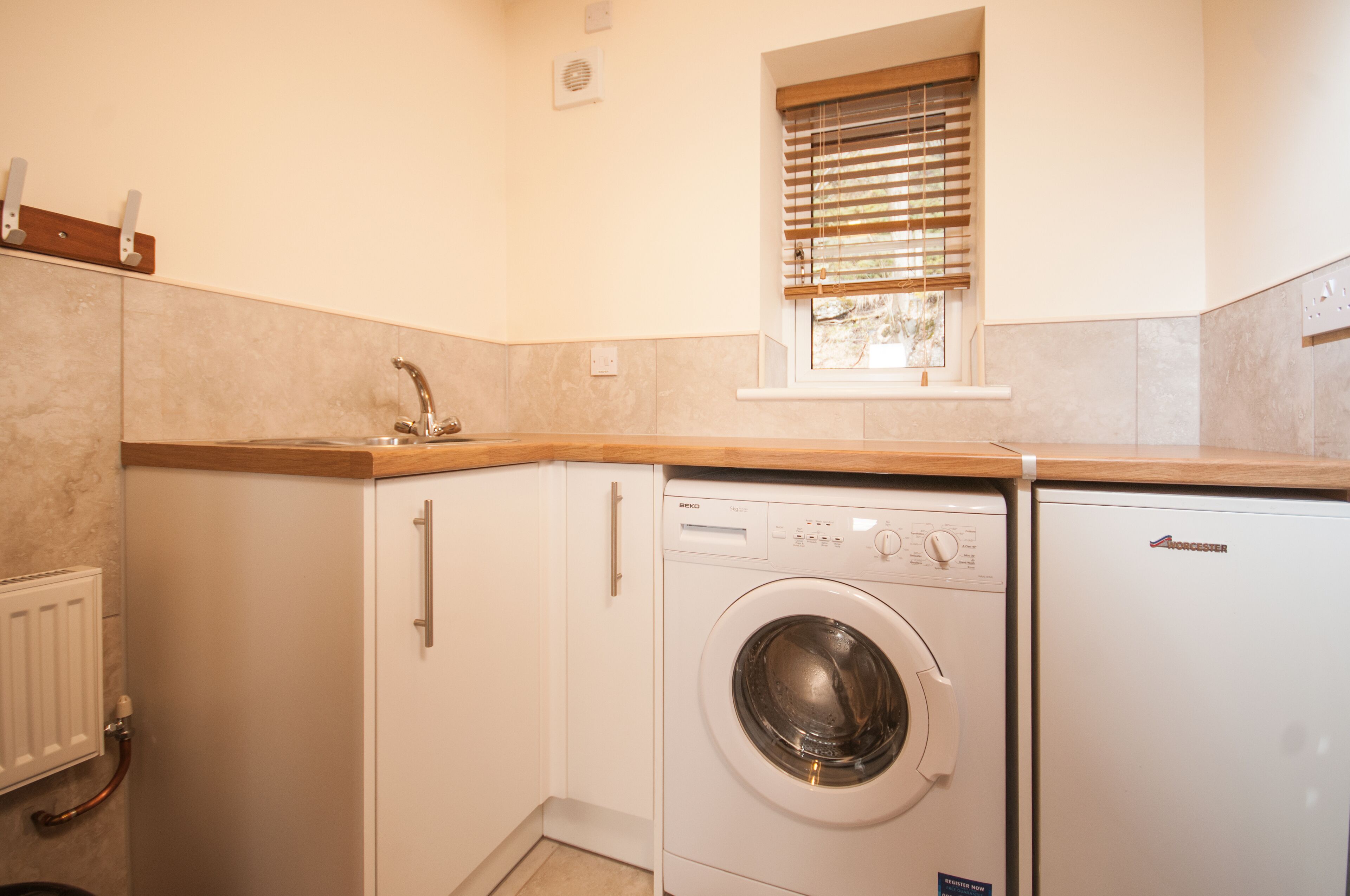 A compact kitchen with a built-in washing machine, a window with blinds, and a radiator.