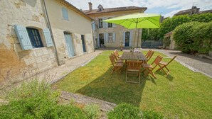 A courtyard with a wooden table and chairs, surrounded by stone buildings with blue shutters.