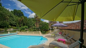 A swimming pool with lounge chairs and a table, surrounded by a garden and a building with a tiled roof.