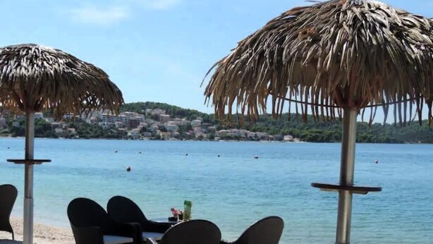 Een tafel met stoelen en een rieten parasol aan het strand, met uitzicht op een kalme zee en een kustplaats op de achtergrond.