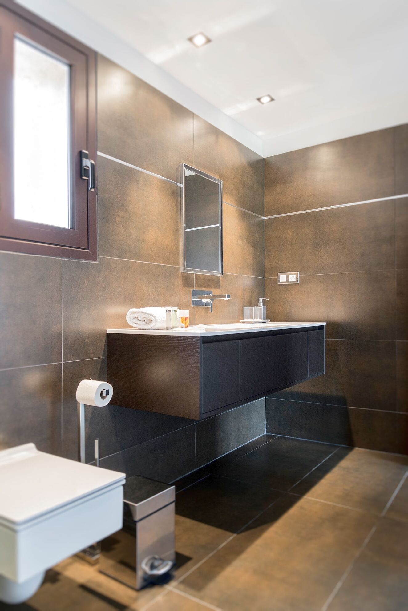 A modern bathroom with a dark brown tiled wall, a large mirror, a white sink, and a toilet.