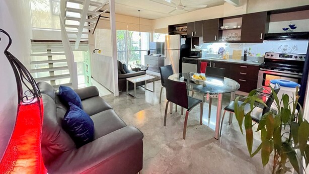 A modern kitchen with dark cabinets, a stainless steel refrigerator, and a built-in oven. There is a dining area with a glass table and chairs, and a sofa with blue cushions. - Spacious 3-Level Loft: Functionality & Comfort for Long-Term Stays (Santa Ana)
