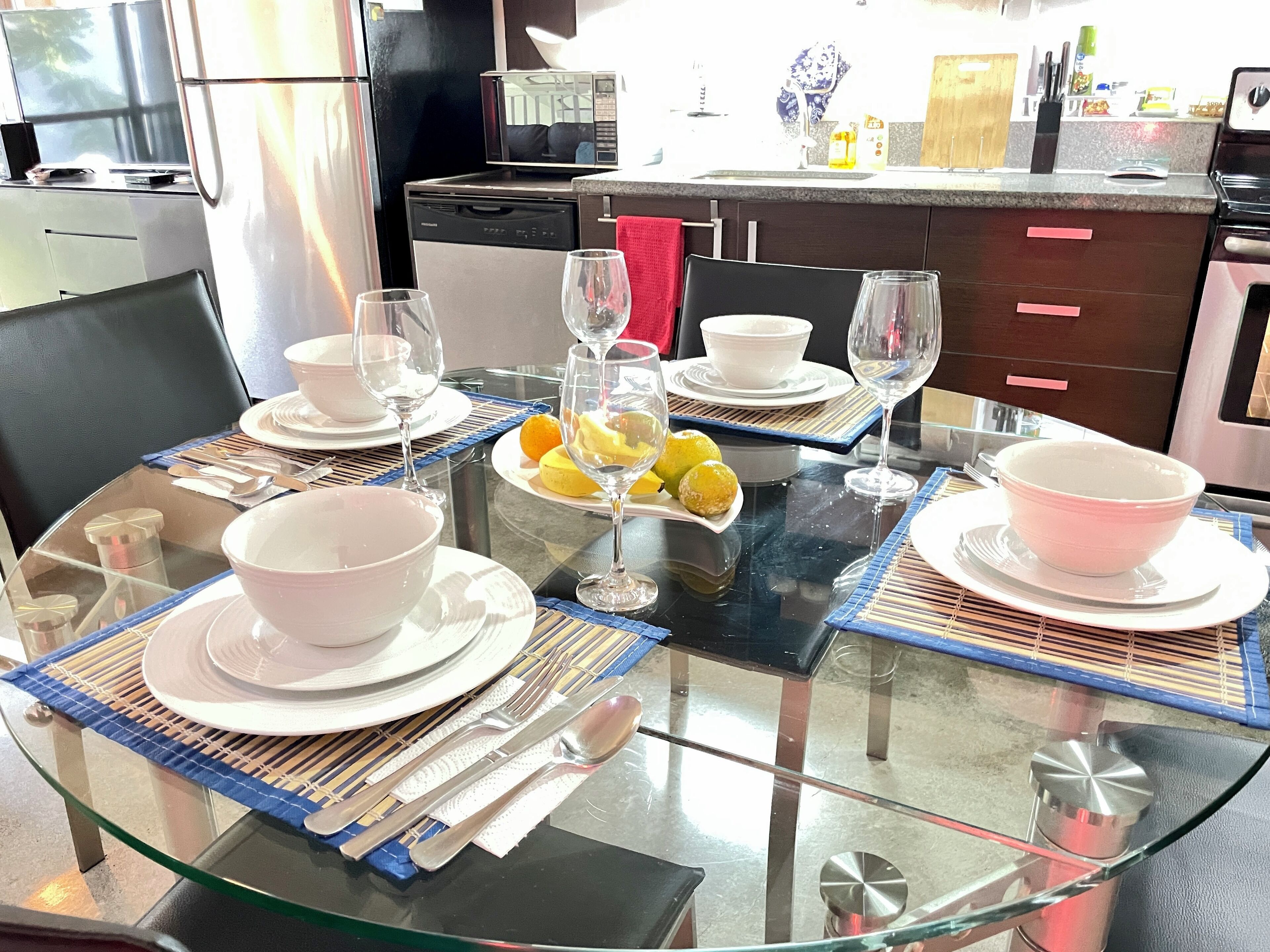Une table à manger dressée avec des bols, des assiettes et des couverts blancs, une table en verre, et une cuisine en arrière-plan.
