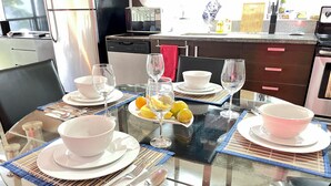 A dining table set with white bowls, plates, and cutlery, a glass table, and a kitchen in the background. - Spacious 3-Level Loft: Functionality & Comfort for Long-Term Stays (Santa Ana)